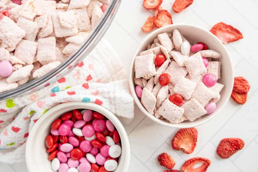 Bowls of pink-coated snack mix with pink, red, and white candies, surrounded by freeze-dried strawberry slices on a patterned cloth.