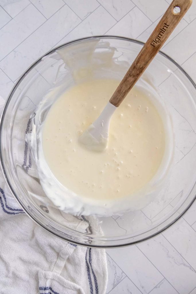 A glass bowl filled with creamy, white batter with a white spatula inside. The bowl sits on a white countertop with a white dish towel with blue stripes.