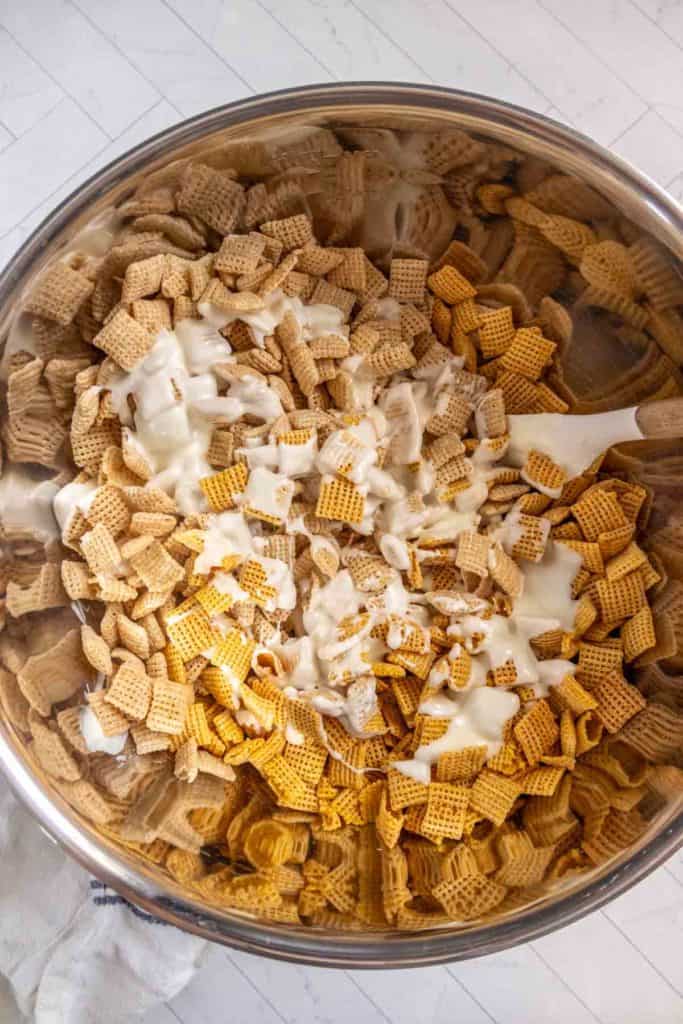 A large metal bowl filled with various types of Chex cereal being mixed with a white coating.