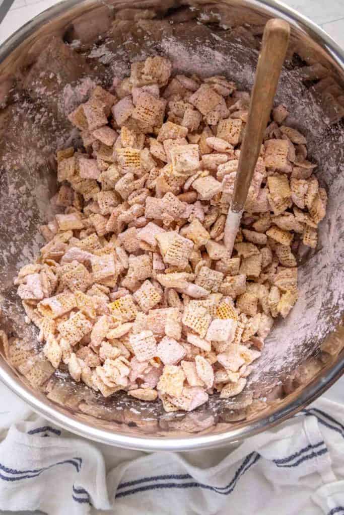 A large metal bowl filled with cereal squares coated with a powdered mixture. A wooden spoon is placed inside the bowl. A striped cloth is partially visible underneath the bowl.