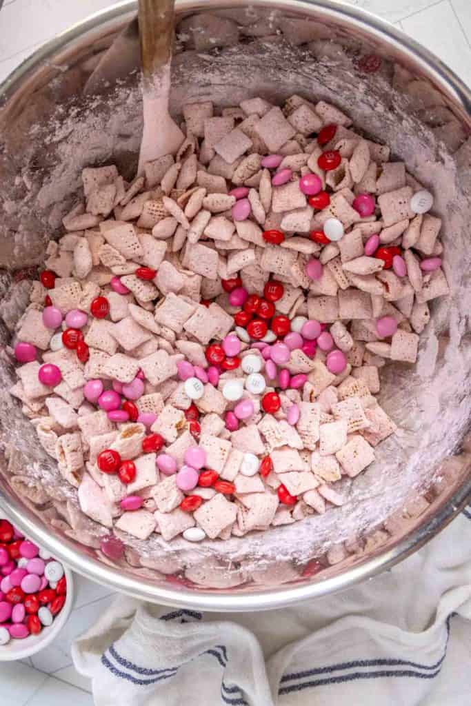 A metal bowl filled with coated cereal pieces and red, pink, and white candy-coated chocolates, with a wooden spoon.