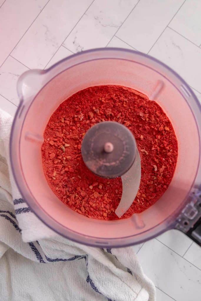 Food processor containing crushed red mixture on a tiled surface, with a striped cloth beside it.