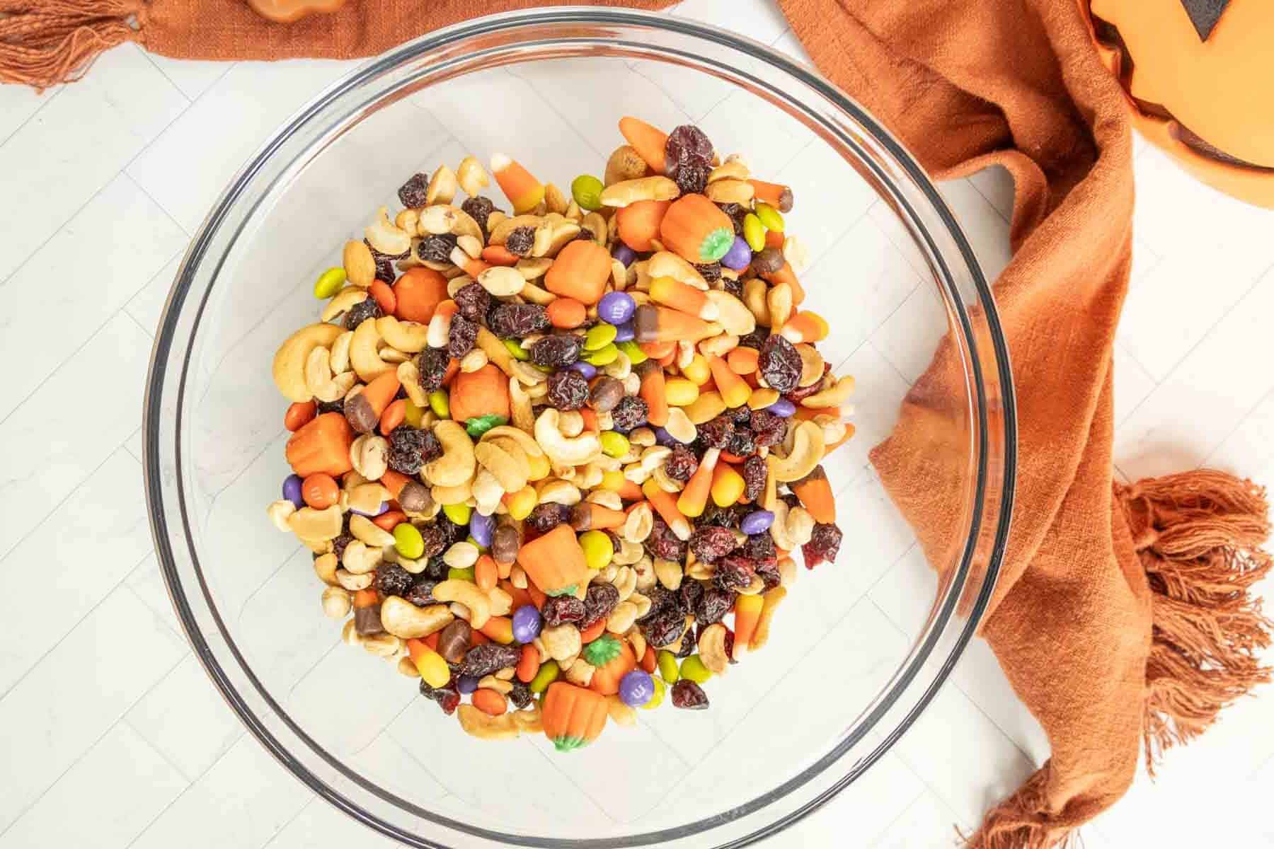 A clear glass bowl filled with a variety of trail mix ingredients, including nuts, raisins, and colorful candy pieces. An orange fabric is placed nearby on a light-colored surface.