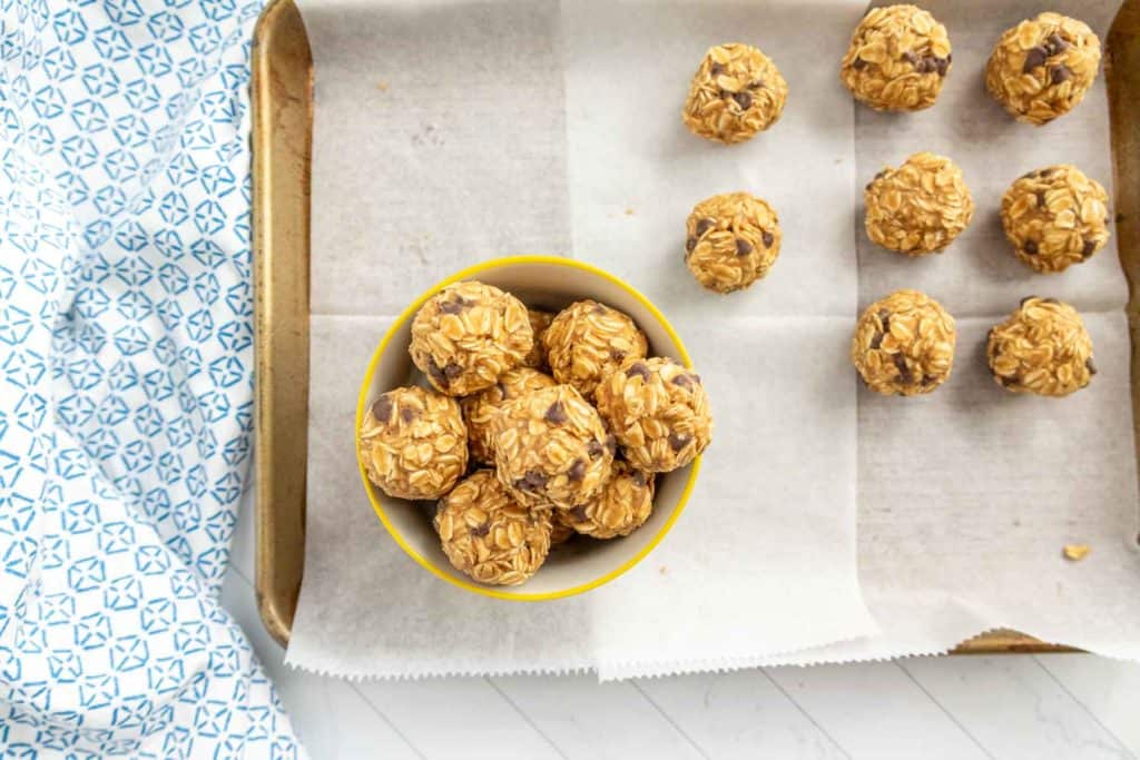 A tray lined with parchment paper holds several peanut butter oatmeal balls, with additional balls in a yellow bowl. A blue patterned cloth is partially visible on the left.