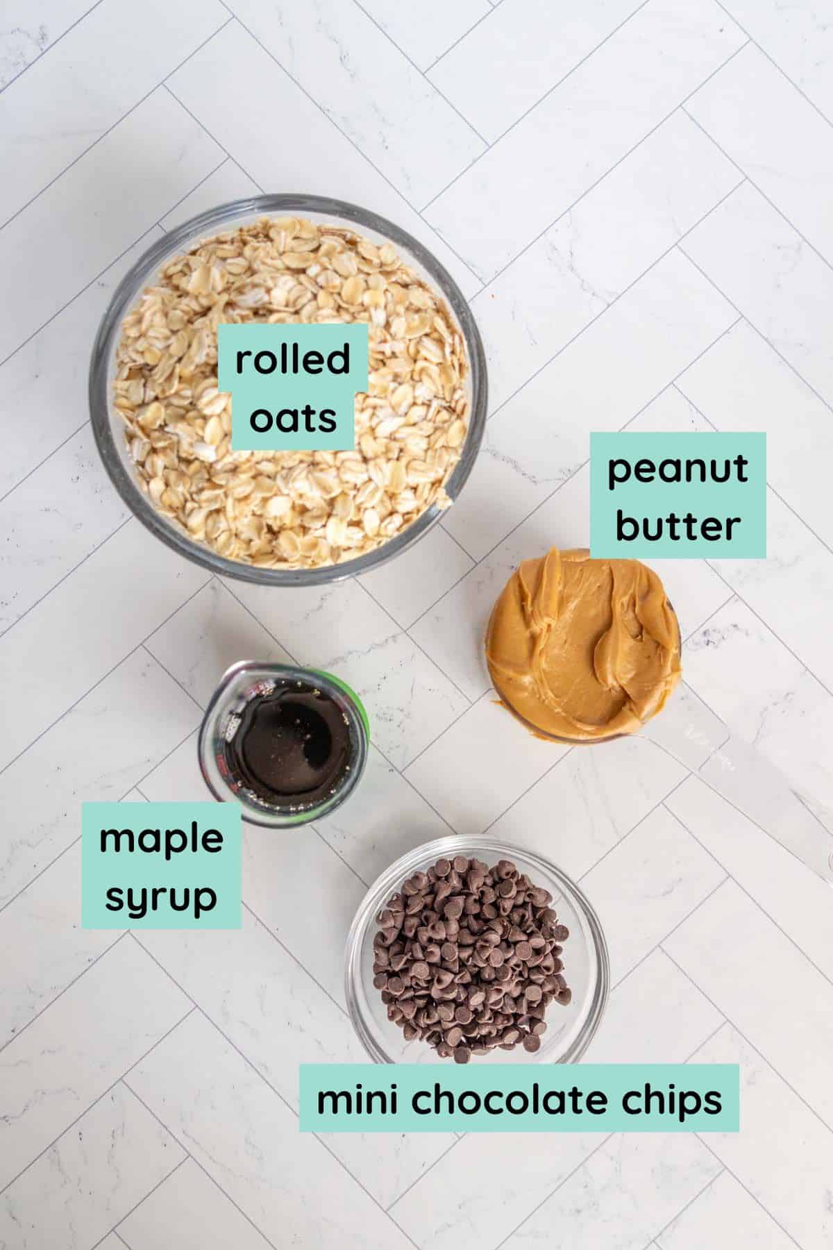 Top-down view of bowls containing rolled oats, peanut butter, maple syrup, and mini chocolate chips, labeled on a white tiled surface.