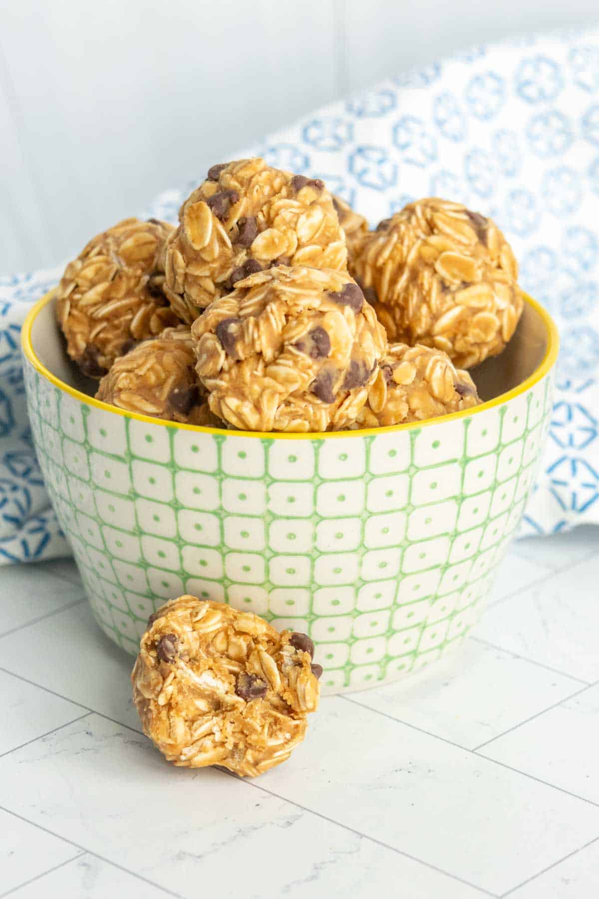 A green checkered bowl filled with oatmeal peanut butter balls, with one ball outside the bowl on a tiled surface.