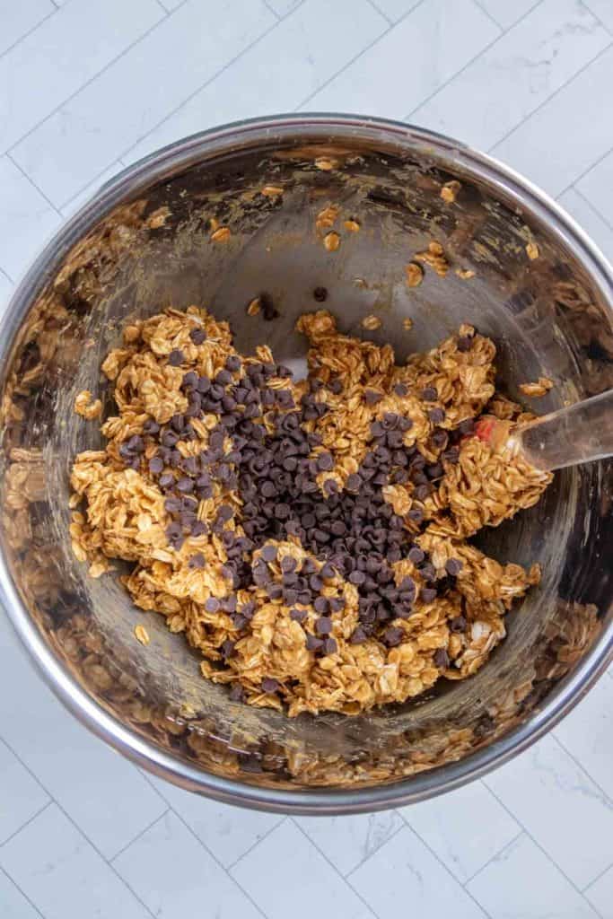 Mixing bowl with oats, peanut butter mixture, and chocolate chips on a tiled surface.