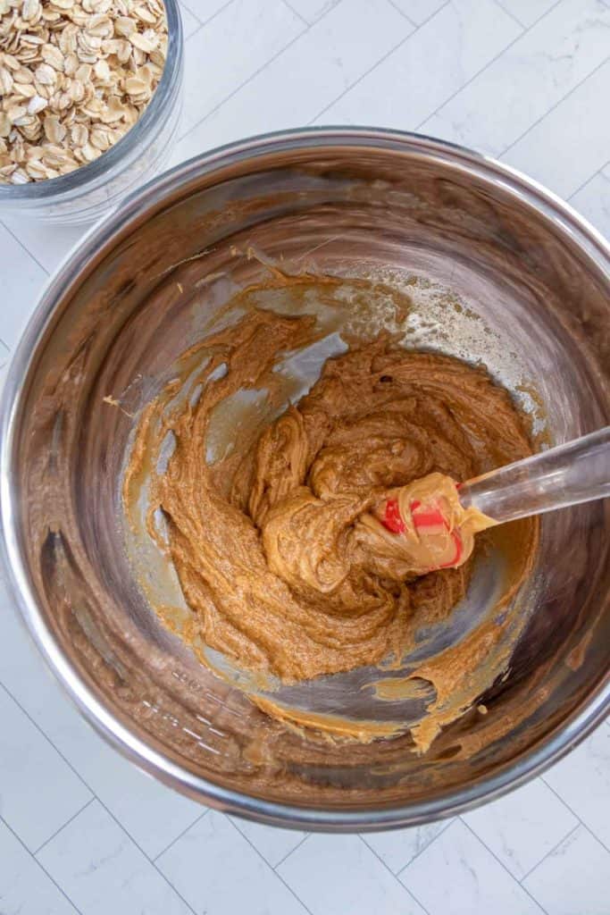 Mixing bowl with a brown batter being stirred by a red spatula. Oats in a separate container are visible nearby on a white tiled surface.