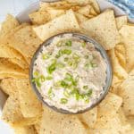 A bowl of creamy dip topped with chopped green onions is surrounded by tortilla chips on a plate.