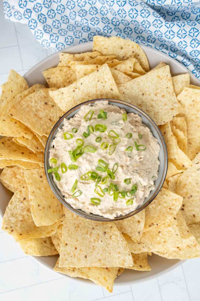 A bowl of creamy dip topped with chopped green onions is surrounded by tortilla chips on a plate.