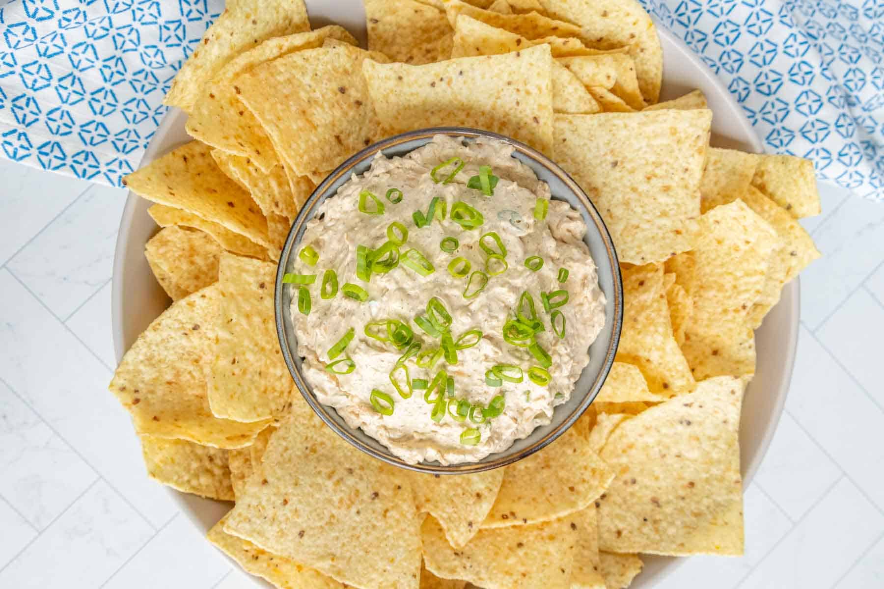 A bowl of dip topped with chopped scallions, surrounded by tortilla chips on a plate.