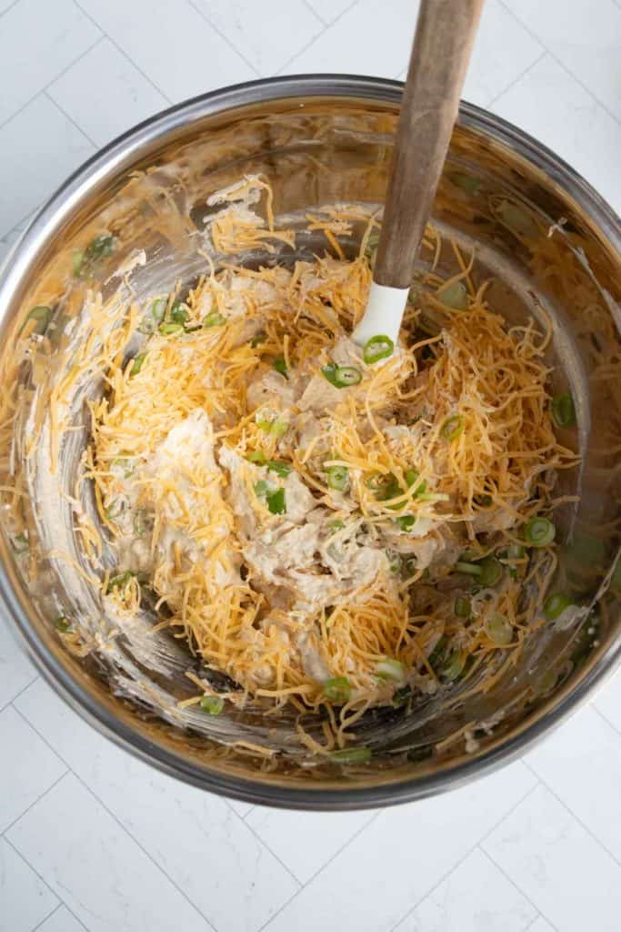 Mixing bowl containing grated cheese, chopped green onions, and a creamy mixture with a spatula on a tiled surface.