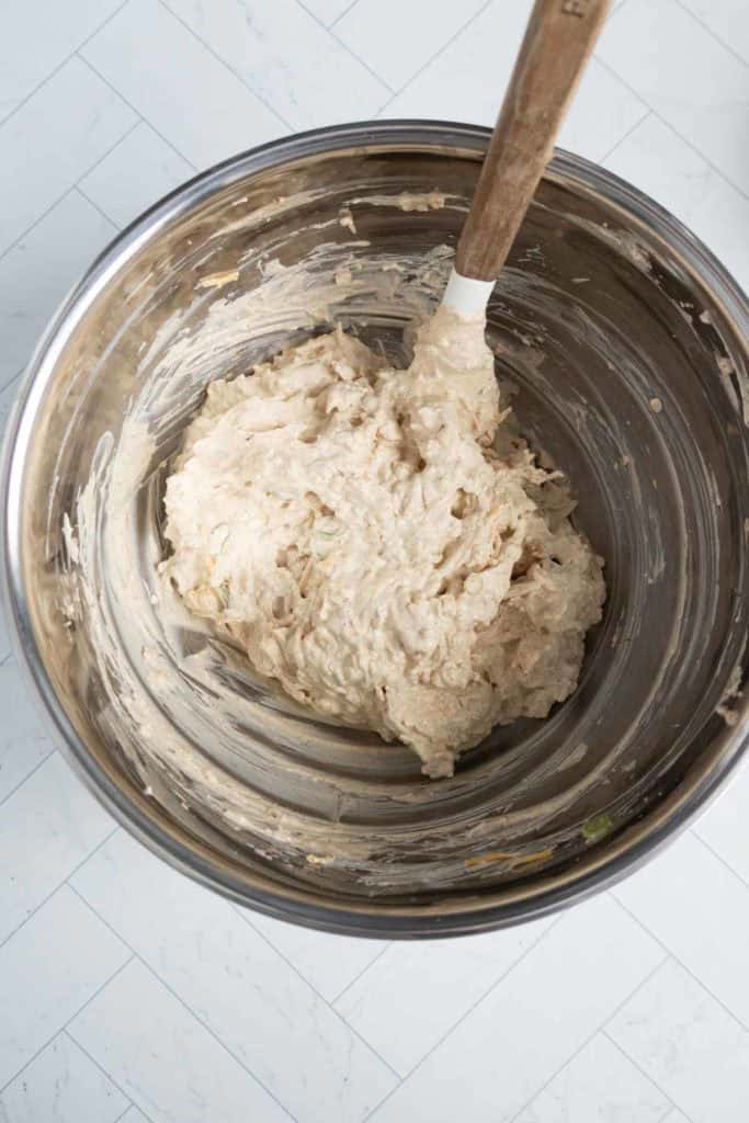 A metal mixing bowl with a wooden spatula contains a thick, light beige dough or batter.