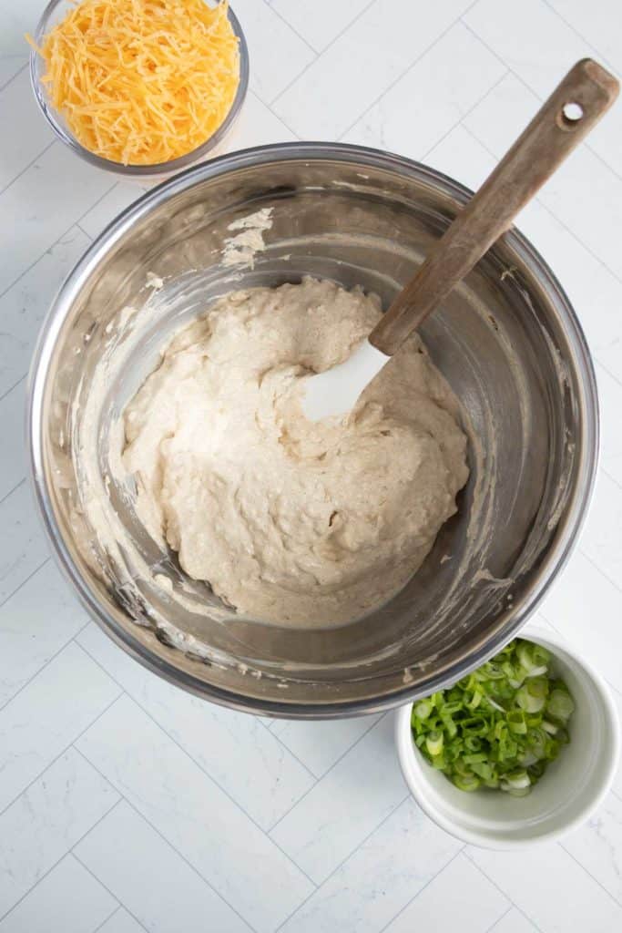 A metal bowl with dough and a spatula, shredded cheese in a small bowl, and chopped green onions in another bowl, placed on a tiled surface.