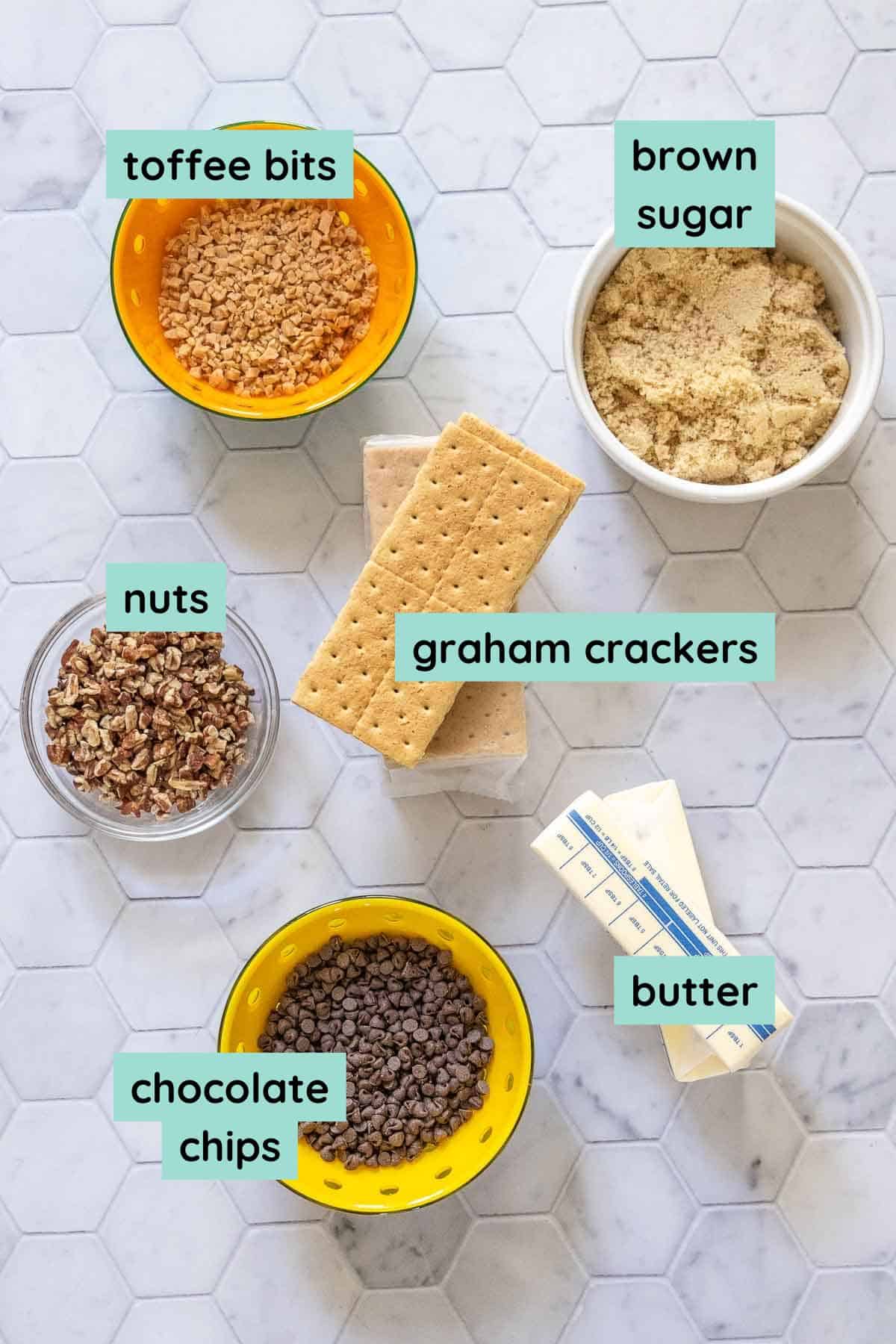 Ingredients on a hexagonal tile surface: toffee bits, brown sugar, nuts, graham crackers, chocolate chips, and butter.