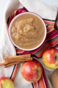 A bowl of applesauce on a striped cloth, surrounded by whole apples and cinnamon sticks.