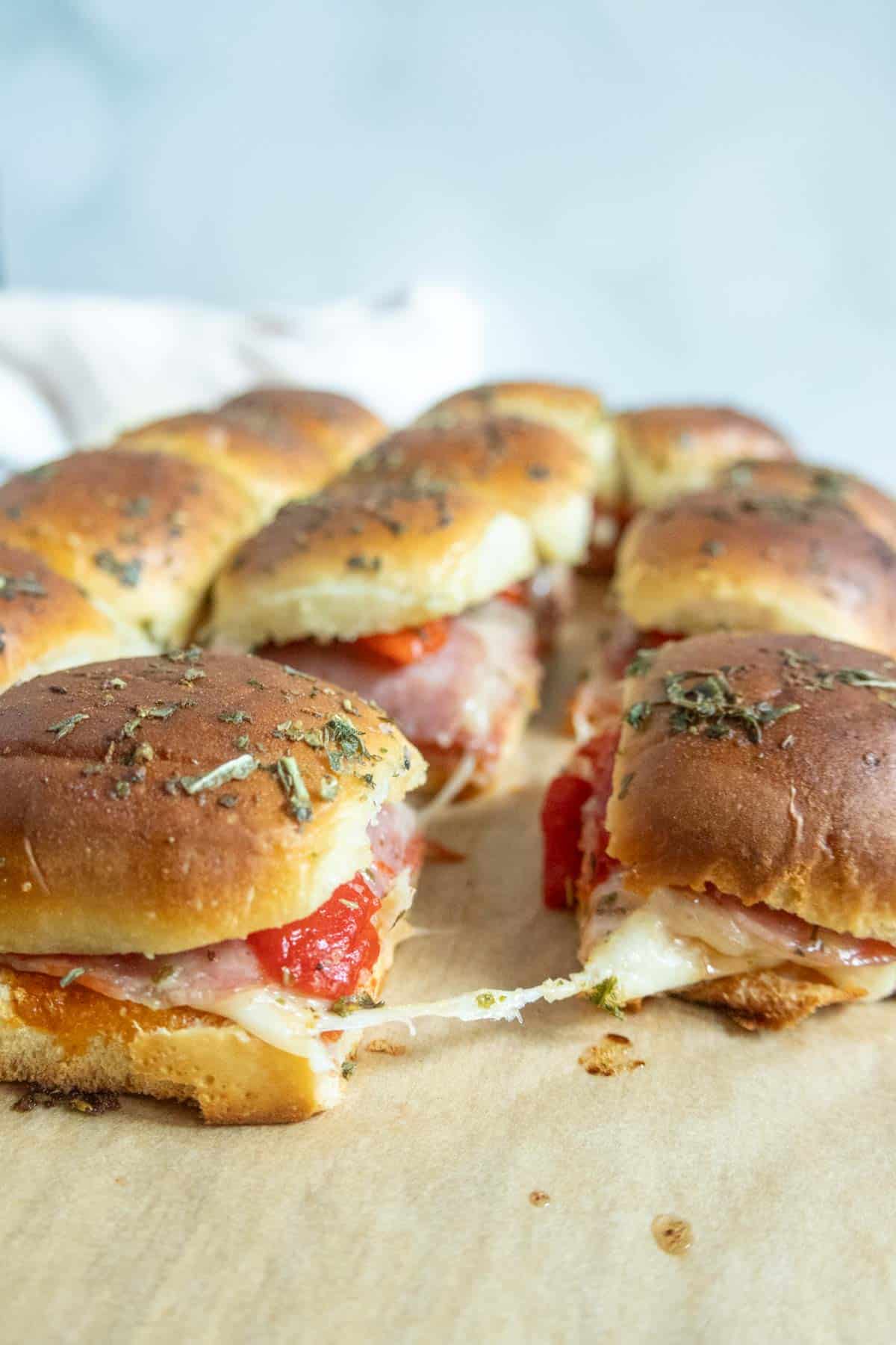 A close-up of baked slider sandwiches filled with melted cheese, ham, and roasted red peppers on parchment paper.