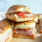 Stacked mini sandwiches with layers of deli meat, cheese, and red peppers on herb-topped bread, arranged on a parchment-lined surface.