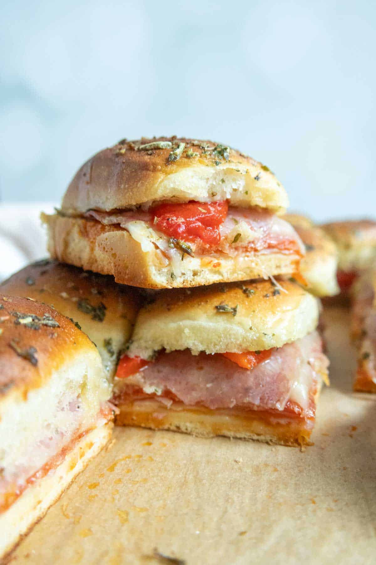 Stacked mini sandwiches with layers of deli meat, cheese, and red peppers on herb-topped bread, arranged on a parchment-lined surface.
