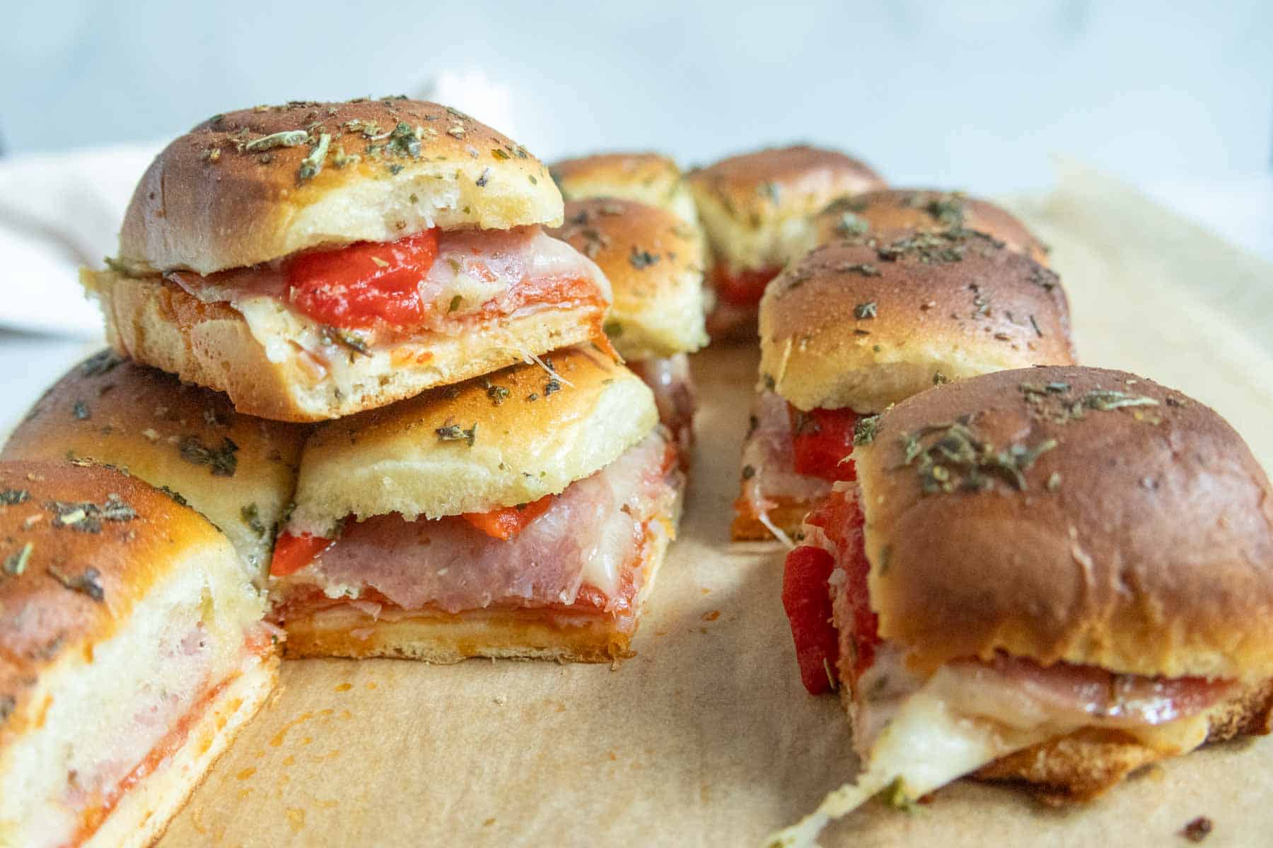 Six slider sandwiches with melted cheese, sliced meats, and red peppers on soft, herb-topped rolls, arranged on a wooden board.