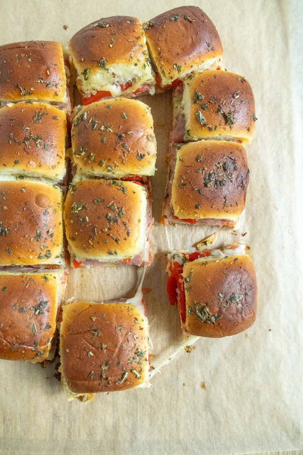 Twelve baked slider sandwiches on parchment paper, with one slider pulled away from the group, showing melted cheese and filling inside.