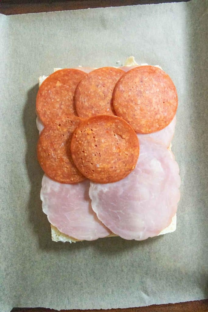 Square slice of bread topped with four slices of ham and five slices of pepperoni on a sheet of parchment paper.