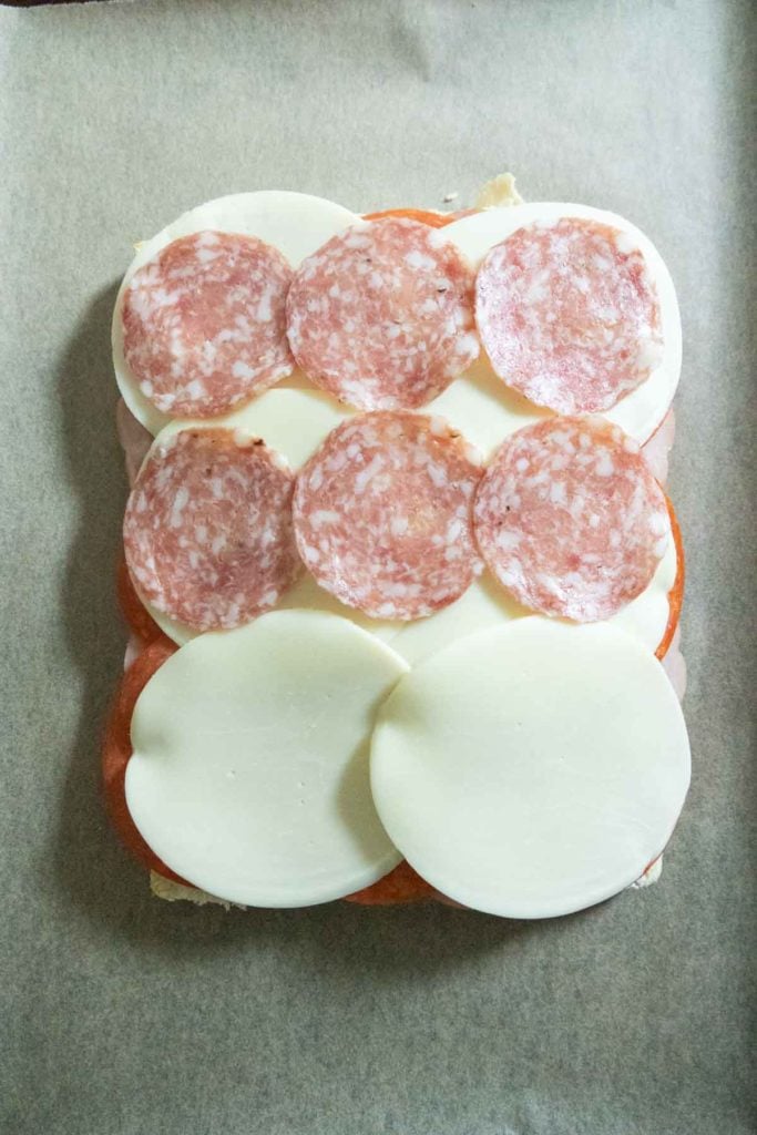 Rectangular sandwich with layers of provolone cheese and round slices of salami on top, placed on parchment paper.