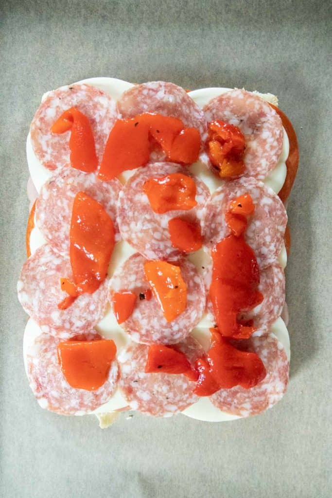 An overhead view of a sandwich topped with slices of salami, mozzarella cheese, and pieces of red peppers on parchment paper.