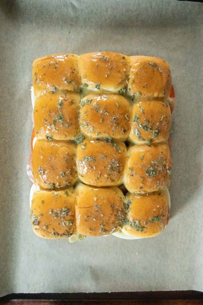 Twelve baked slider rolls brushed with herb butter are arranged closely together on a sheet of parchment paper.