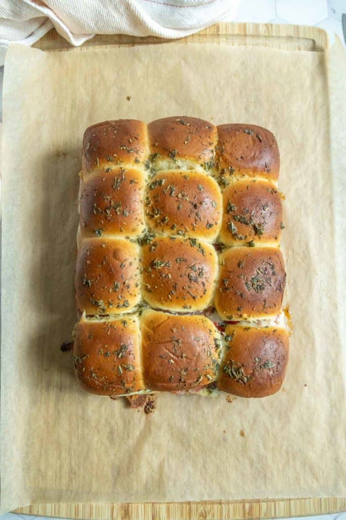 A rectangular batch of golden brown bread rolls topped with herbs, placed on a sheet of parchment paper on a wooden surface.
