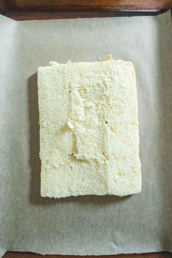 A rectangular block of white bread with a rough surface sits on a sheet of parchment paper.