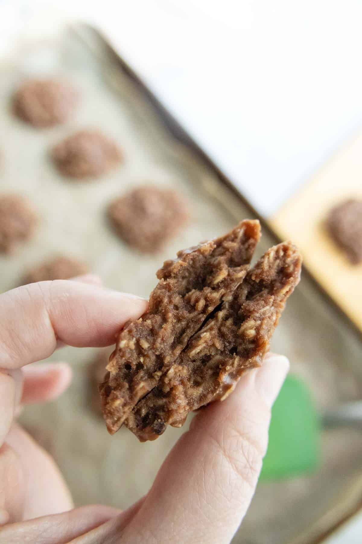 A hand holds a split chocolate oatmeal cookie, revealing its chewy texture inside, with more cookies on a parchment-lined baking sheet in the background.