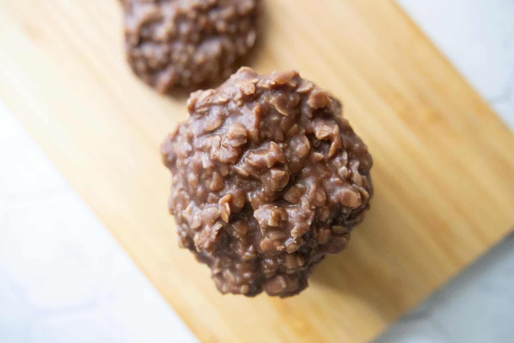A no-bake chocolate oatmeal cookie sits on a wooden cutting board with another cookie partially visible in the background.