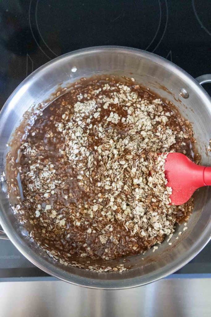 A saucepan with oats and chocolate mixture being stirred with a red spatula on a stovetop.