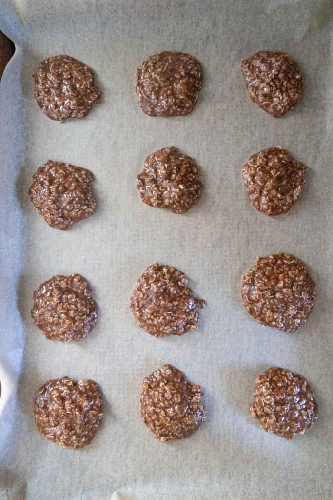 Twelve chocolate oatmeal cookies are arranged in three rows on a parchment-lined baking sheet.