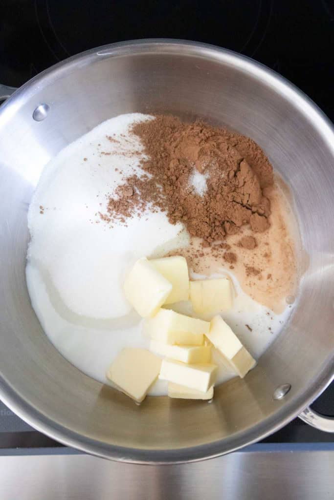 Stainless steel pan containing granulated sugar, cocoa powder, butter, and milk, prepared for mixing on a stovetop.