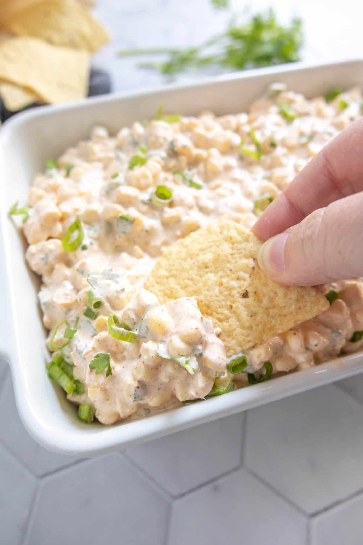 A hand dips a tortilla chip into a creamy, chunky dip topped with chopped green onions in a white dish.