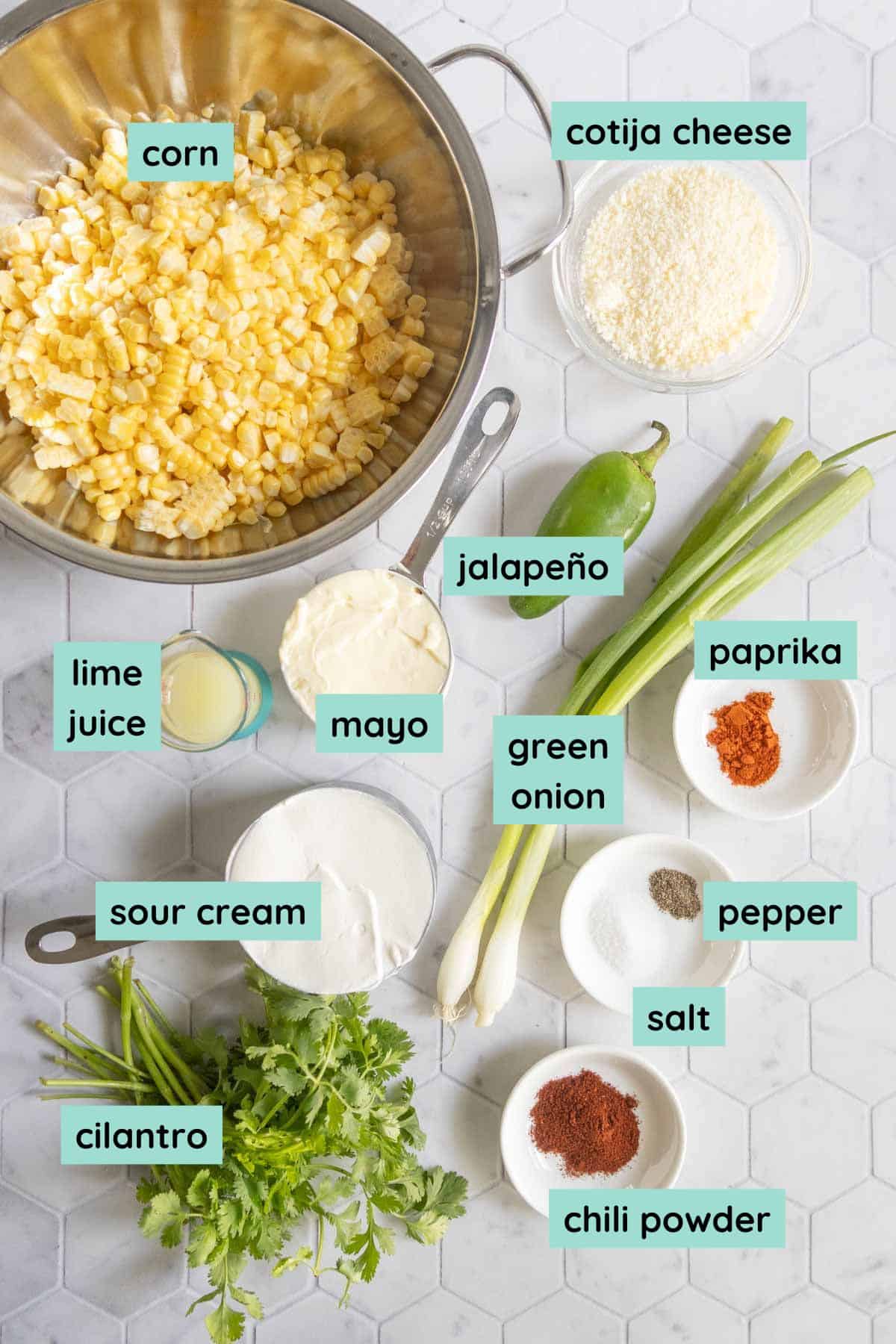Overhead view of ingredients for a corn dish, including corn, cotija cheese, jalapeño, lime juice, mayo, green onion, paprika, sour cream, pepper, salt, chili powder, and cilantro.