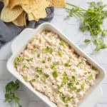 A white dish filled with creamy corn dip is garnished with chopped green onions, surrounded by tortilla chips and fresh cilantro on a white hexagon-tiled surface.
