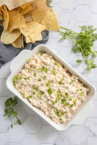 A white dish filled with creamy corn dip is garnished with chopped green onions, surrounded by tortilla chips and fresh cilantro on a white hexagon-tiled surface.