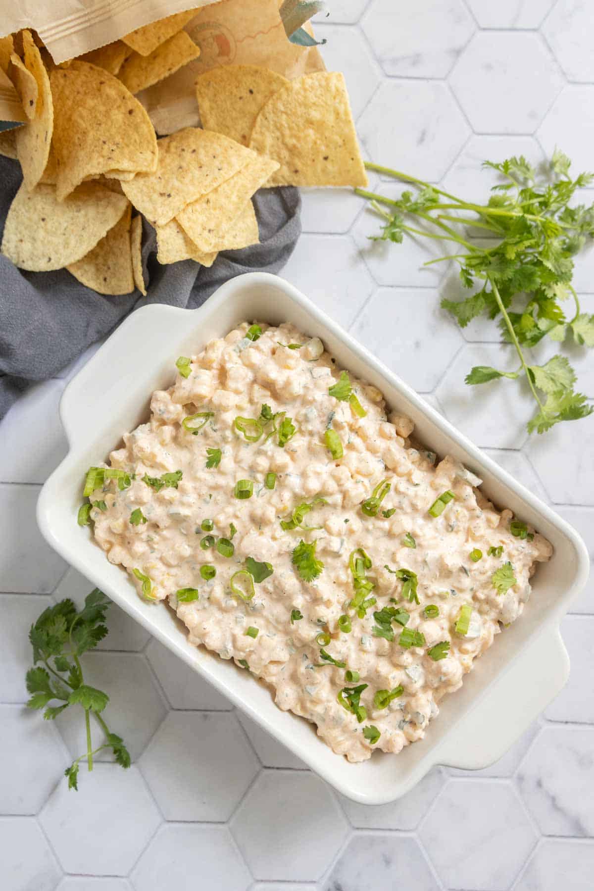 A white dish filled with creamy corn dip is garnished with chopped green onions, surrounded by tortilla chips and fresh cilantro on a white hexagon-tiled surface.