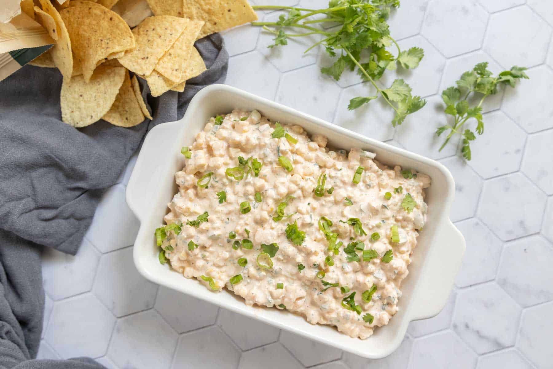 A white dish filled with creamy dip, garnished with chopped green herbs, sits on a white hexagon-tiled surface next to tortilla chips and fresh cilantro.