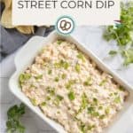 A white dish filled with creamy street corn dip, garnished with chopped green herbs, sits on a white surface next to fresh cilantro and tortilla chips.