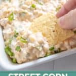 A hand dips a tortilla chip into a creamy street corn dip garnished with chopped green onions in a white dish.