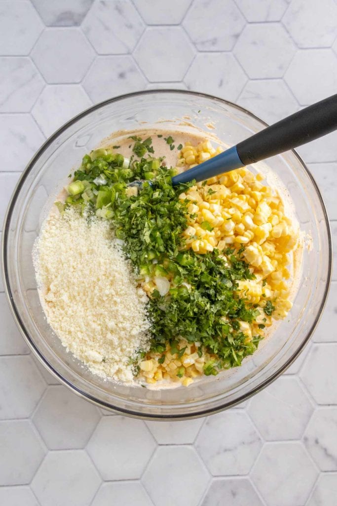 A glass bowl contains chopped green herbs, corn kernels, grated cheese, and batter, with a black spatula resting inside. The bowl is on a hexagonal tile countertop.