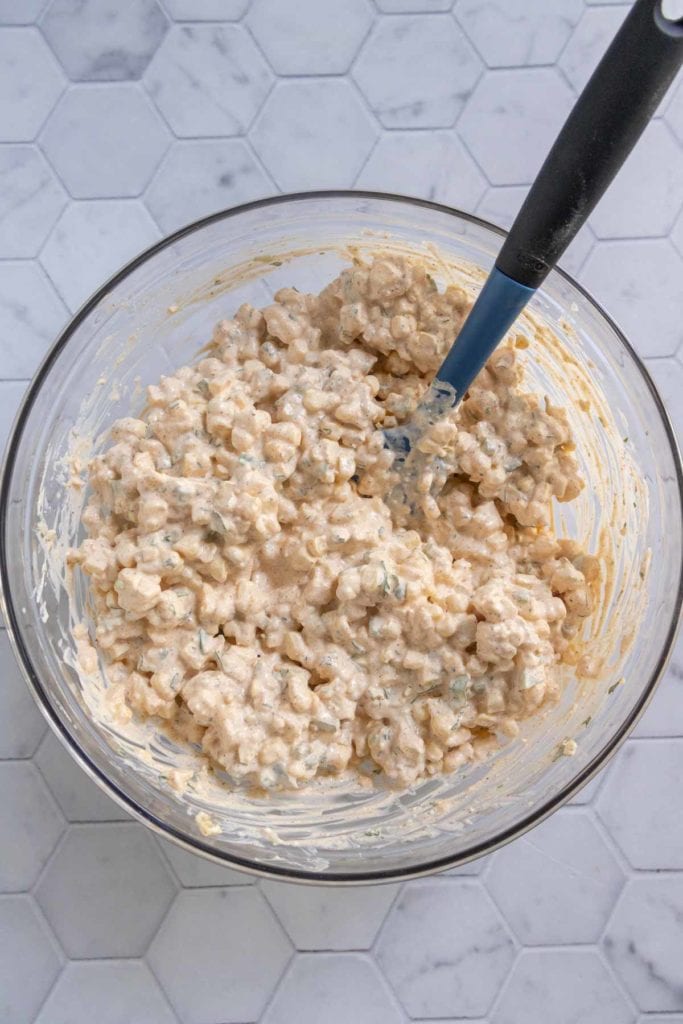 A glass bowl filled with a creamy rice salad mixture, with a black spoon resting inside, placed on a white hexagon tile surface.
