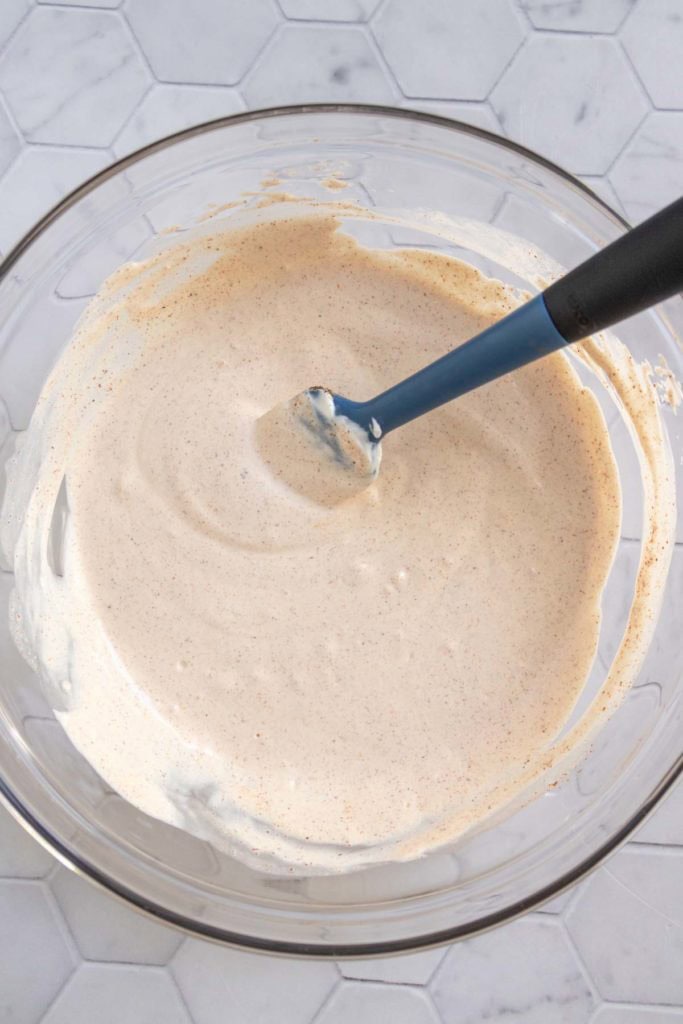 A glass bowl containing a light brown, creamy mixture being stirred with a blue spatula, placed on a hexagonal tile surface.