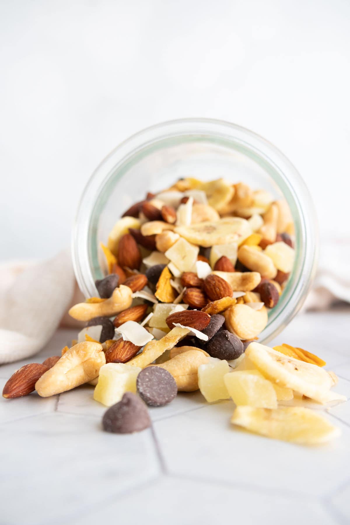 A glass jar tipped over, spilling mixed nuts, dried fruit pieces, and chocolate chips onto a light surface.