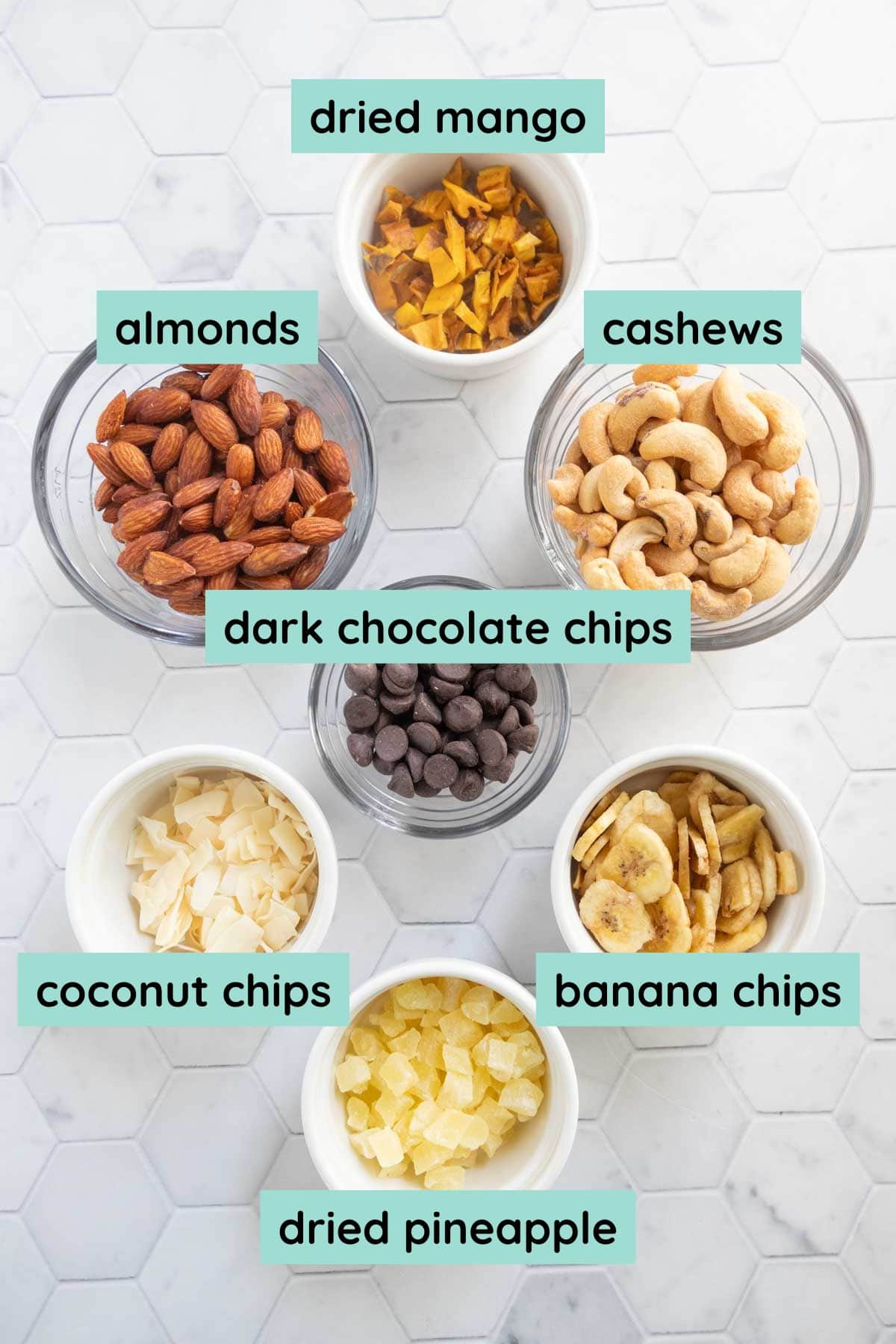 Seven bowls containing dried mango, cashews, banana chips, dried pineapple, coconut chips, dark chocolate chips, and almonds, arranged on a white tiled surface.
