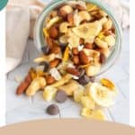 A glass jar tipped over, spilling tropical trail mix containing banana chips, chocolate chips, almonds, cashews, coconut flakes, and dried fruit onto a marble surface.